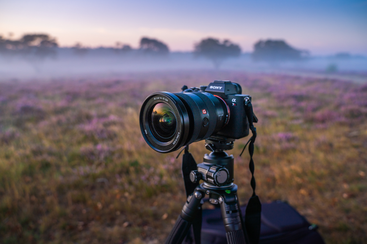  A Sony Alpha 7R III camera with a wide-angle lens attached is sitting on a tripod in a field of purple flowers with a beautiful sunrise in the background.