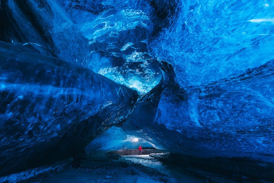 A woman in red stands in the background of an ice cave - Iceland Photography | Everything You Need To Know
