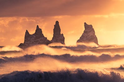 The Reynisdrangar sea stacks look spectacular in the ocean under a yellow-orange sky that reflects on the surrounding waters.