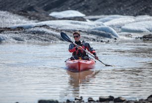A person paddles across the calm waters of the Solheimajokull glacier lagoon with icebergs in the background.